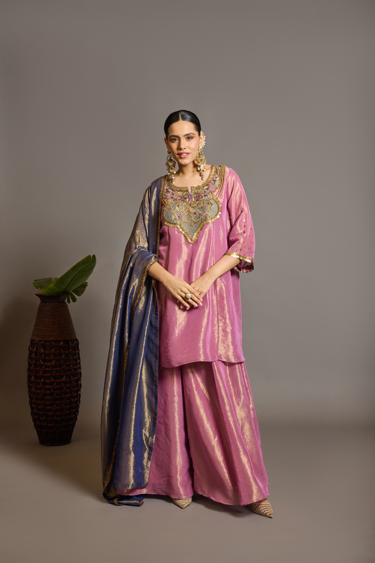 A luminous rose-pink kurta and flared palazzo set crafted in rich,metallic-finish fabric. Featuring an ornate multicolour embroidered neckline with intricate zardozi inspired detailing. Paired with a contrasting deep-blue dupatta finished with elegant gold borders. A festive ensemble that beautifully blends contemporary ease with traditional opulence. Shop Now!