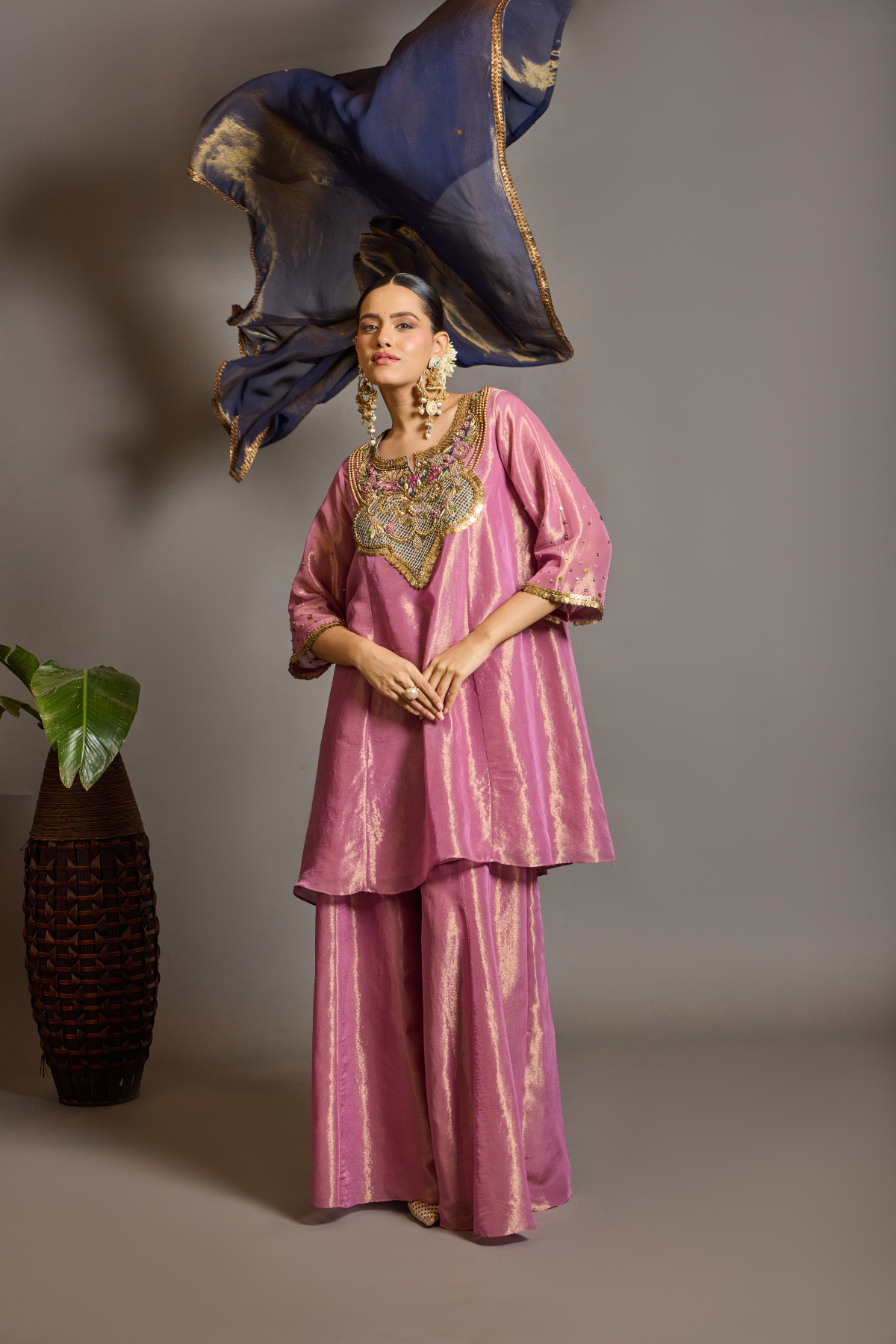 A luminous rose-pink kurta and flared palazzo set crafted in rich,metallic-finish fabric. Featuring an ornate multicolour embroidered neckline with intricate zardozi inspired detailing. Paired with a contrasting deep-blue dupatta finished with elegant gold borders. A festive ensemble that beautifully blends contemporary ease with traditional opulence. Shop Now!
