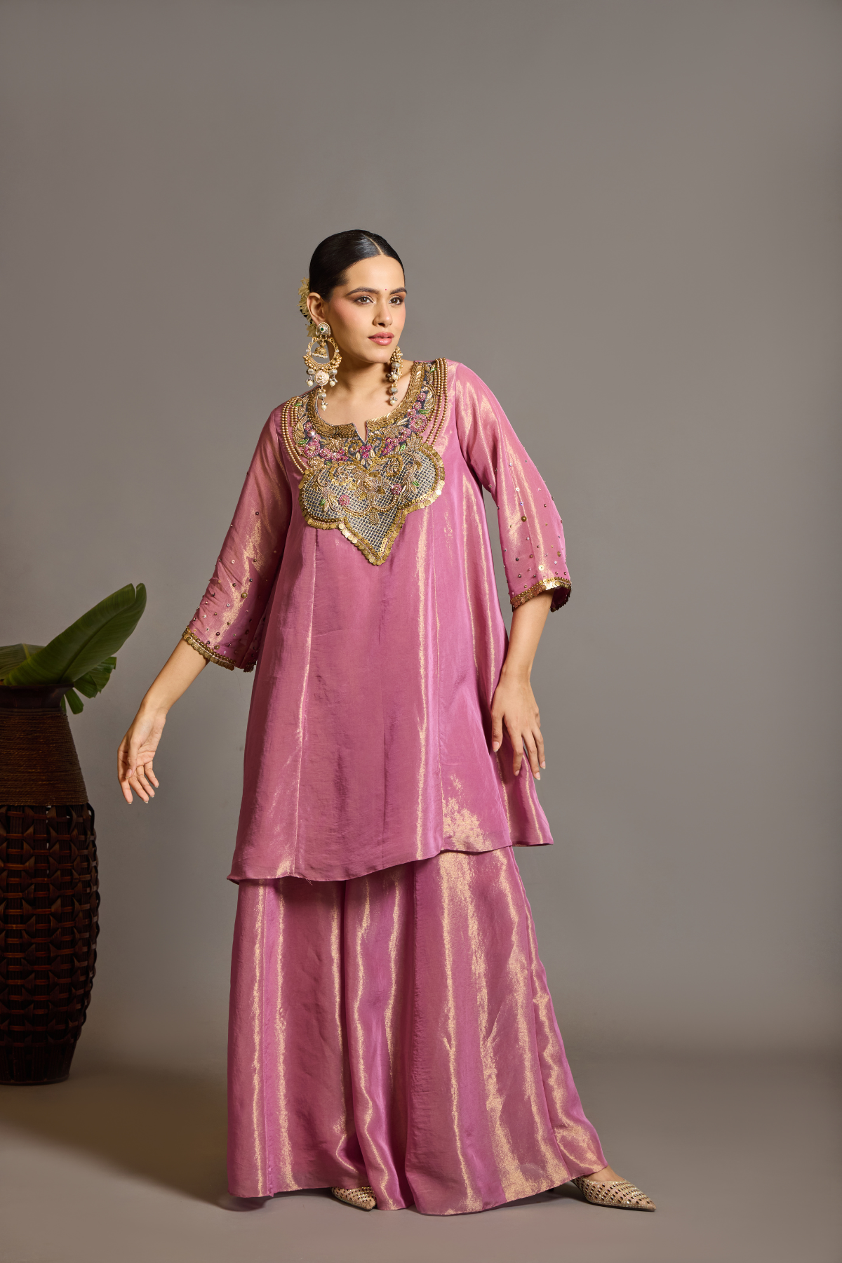 A luminous rose-pink kurta and flared palazzo set crafted in rich,metallic-finish fabric. Featuring an ornate multicolour embroidered neckline with intricate zardozi inspired detailing. Paired with a contrasting deep-blue dupatta finished with elegant gold borders. A festive ensemble that beautifully blends contemporary ease with traditional opulence. Shop Now!