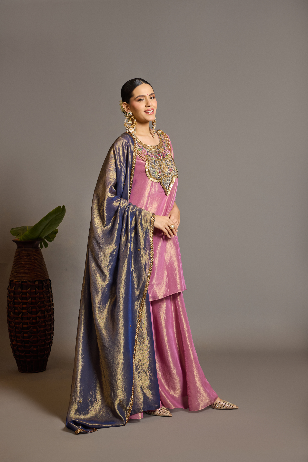 A luminous rose-pink kurta and flared palazzo set crafted in rich,metallic-finish fabric. Featuring an ornate multicolour embroidered neckline with intricate zardozi inspired detailing. Paired with a contrasting deep-blue dupatta finished with elegant gold borders. A festive ensemble that beautifully blends contemporary ease with traditional opulence. Shop Now!