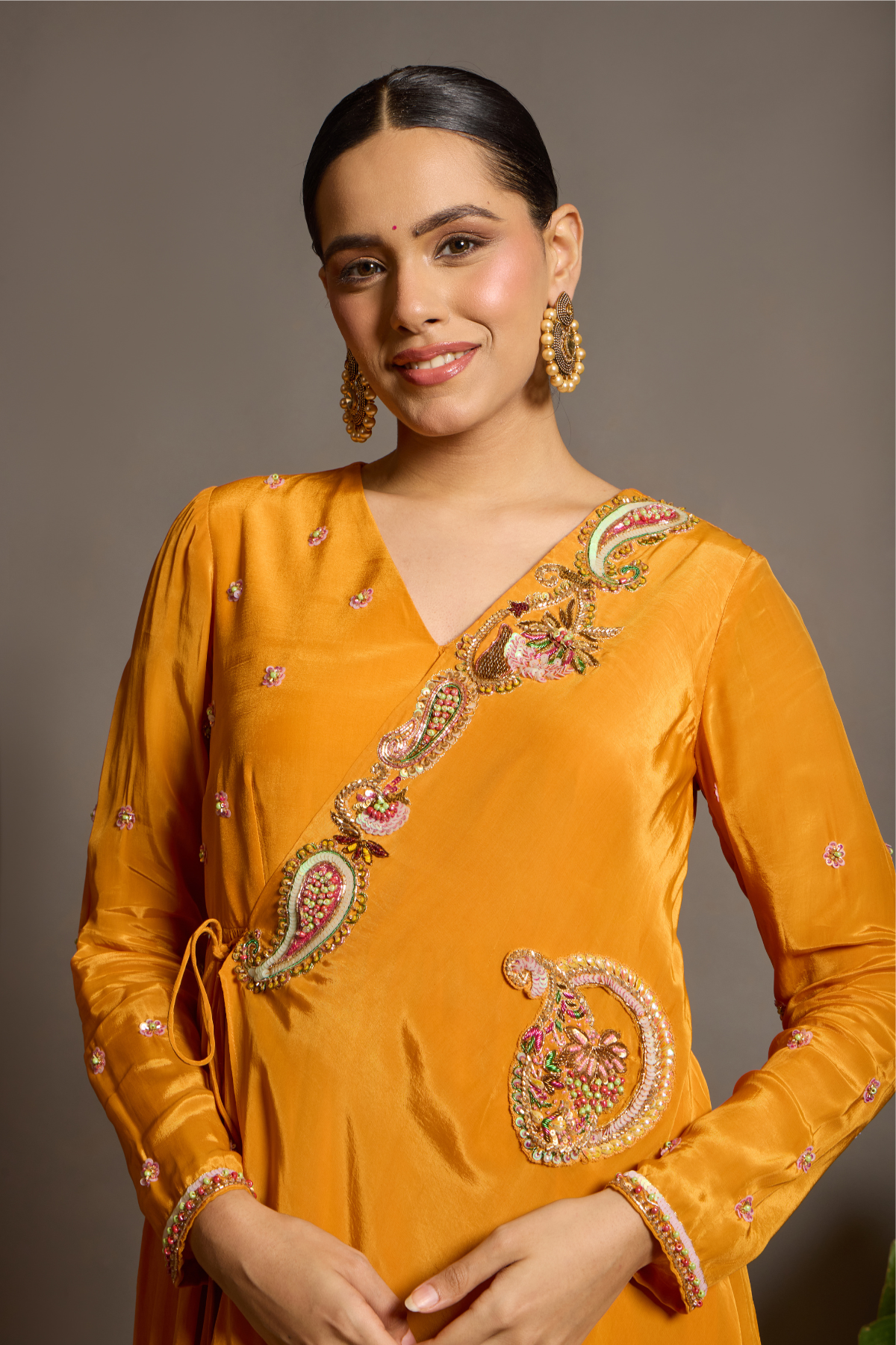 A vibrant marigold-orange Anarkali crafted in a fluid, lustrous fabric with a graceful layered silhouette. Adorned with intricate multicolour paisley embroidery and delicate scattered embellishments. Features a flattering tie-up waist with handcrafted tassels and elegant embroidered work. Shop Now!