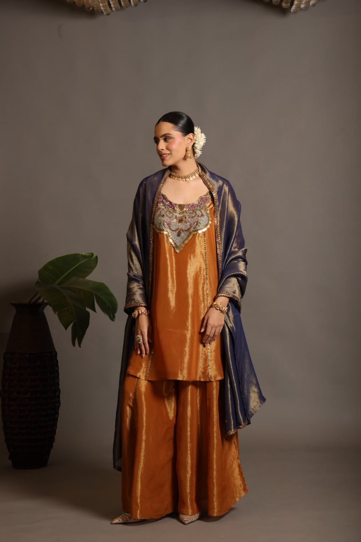A luminous burnt-orange kurta in a rich metallic finish, elevated with an ornate handcrafted embroidered yoke. The statement neckline features intricate multicolour florals, paisleys,sequins and antique-gold detailing. Layered with a deep-navy blue metallic dupatta finished with ornate borders and delicate embellishment. A festive look that blends contemporary glamour with traditional Indian craftsmanship. Shop Now!