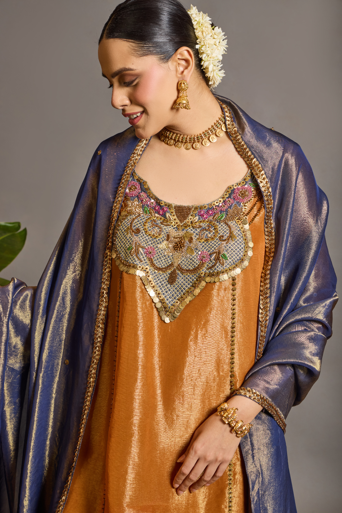 A luminous burnt-orange kurta in a rich metallic finish, elevated with an ornate handcrafted embroidered yoke. The statement neckline features intricate multicolour florals, paisleys,sequins and antique-gold detailing. Layered with a deep-navy blue metallic dupatta finished with ornate borders and delicate embellishment. A festive look that blends contemporary glamour with traditional Indian craftsmanship. Shop Now!