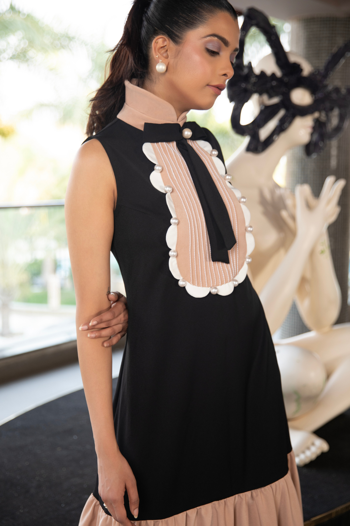 The dress features a structured black bodice with a distinctive scalloped pleated bib detail in beige on the chest finished with a black bow and pearl button Shop Now!