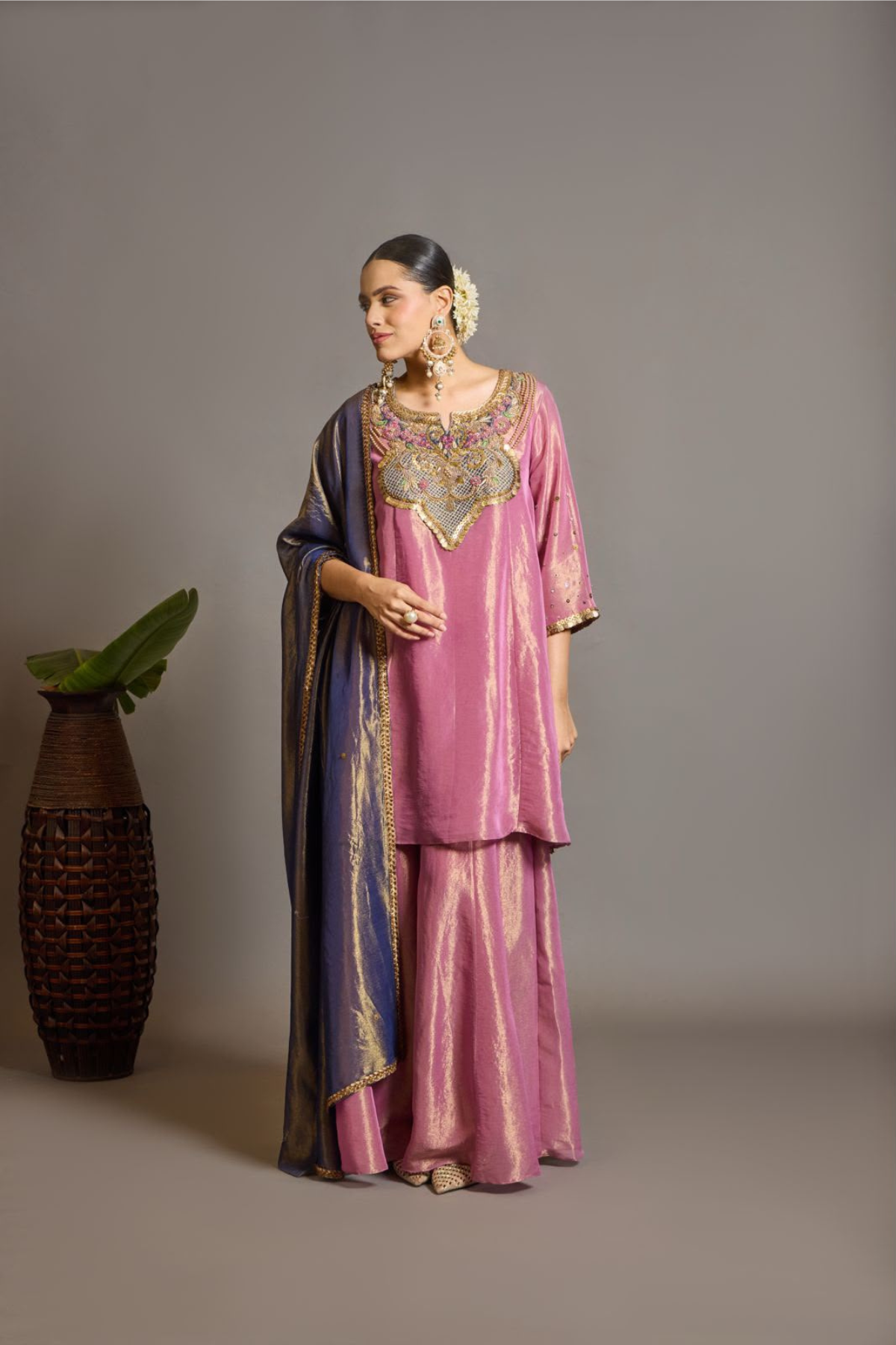 A luminous rose-pink kurta and flared palazzo set crafted in rich,metallic-finish fabric. Featuring an ornate multicolour embroidered neckline with intricate zardozi inspired detailing. Paired with a contrasting deep-blue dupatta finished with elegant gold borders. A festive ensemble that beautifully blends contemporary ease with traditional opulence. Shop Now!