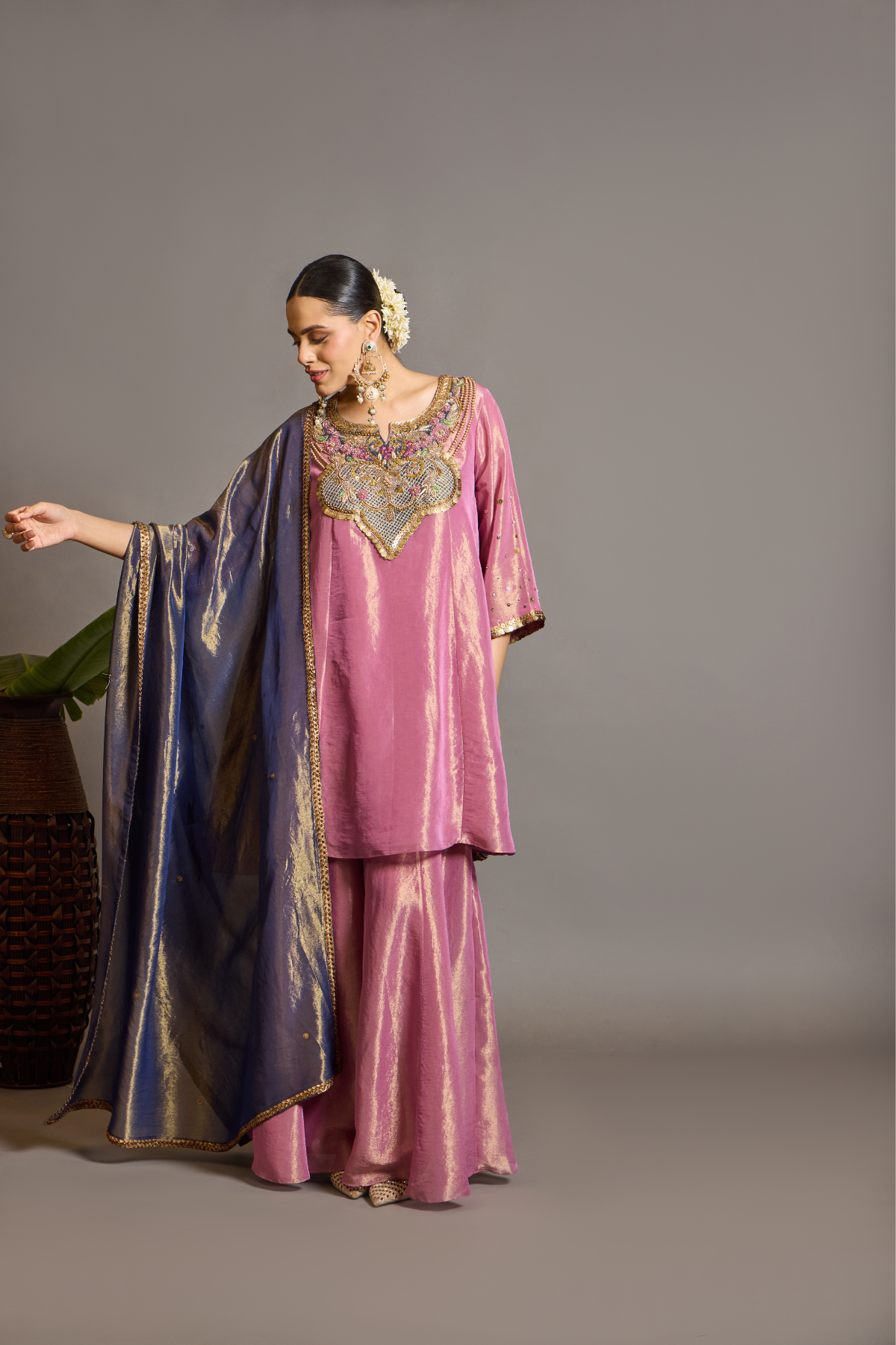 A luminous rose-pink kurta and flared palazzo set crafted in rich,metallic-finish fabric. Featuring an ornate multicolour embroidered neckline with intricate zardozi inspired detailing. Paired with a contrasting deep-blue dupatta finished with elegant gold borders. A festive ensemble that beautifully blends contemporary ease with traditional opulence. Shop Now!
