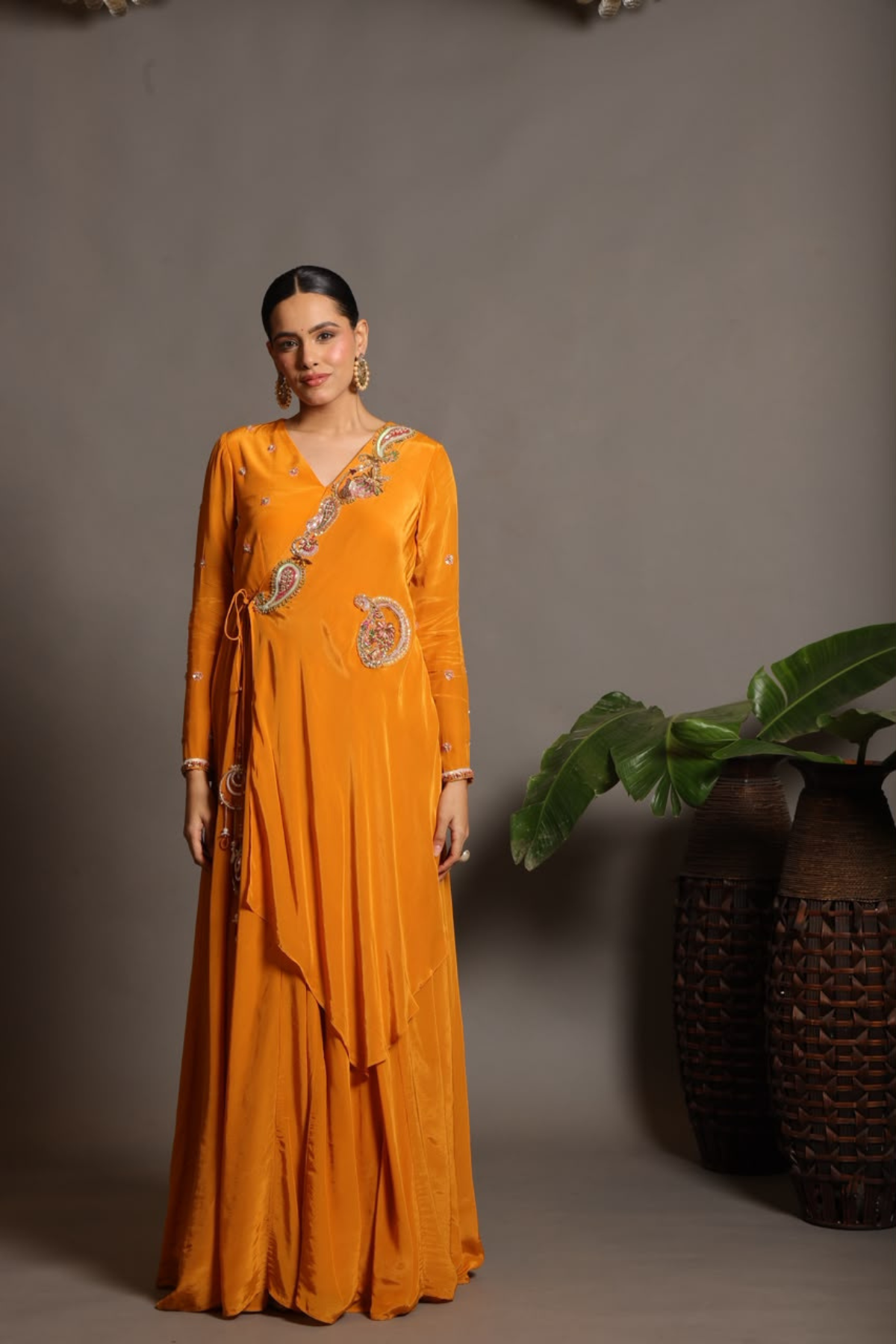 A vibrant marigold-orange Anarkali crafted in a fluid, lustrous fabric with a graceful layered silhouette. Adorned with intricate multicolour paisley embroidery and delicate scattered embellishments. Features a flattering tie-up waist with handcrafted tassels and elegant embroidered work. Shop Now!