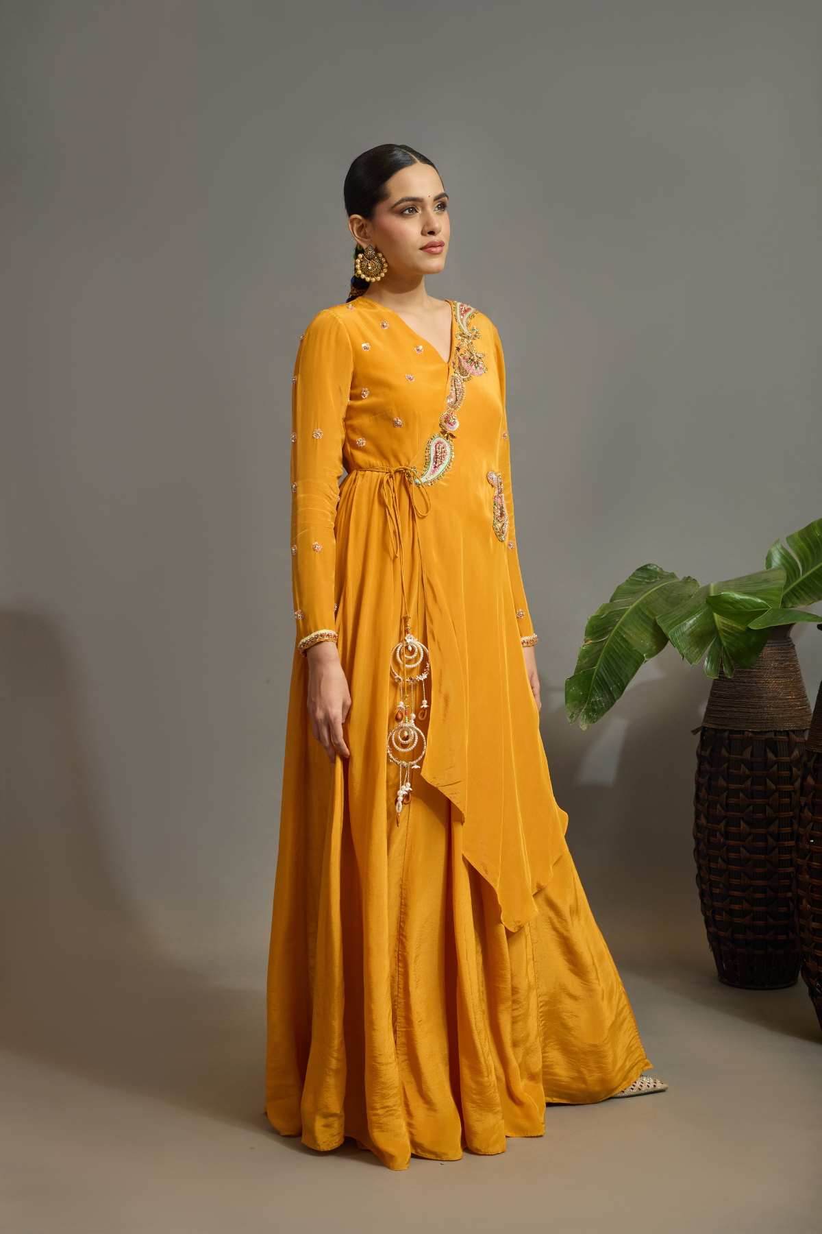 A vibrant marigold-orange Anarkali crafted in a fluid, lustrous fabric with a graceful layered silhouette. Adorned with intricate multicolour paisley embroidery and delicate scattered embellishments. Features a flattering tie-up waist with handcrafted tassels and elegant embroidered work. Shop Now!
