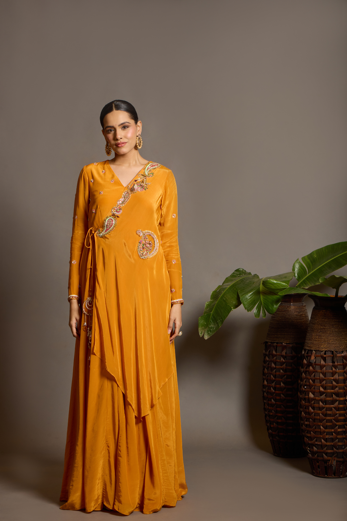 A vibrant marigold-orange Anarkali crafted in a fluid, lustrous fabric with a graceful layered silhouette. Adorned with intricate multicolour paisley embroidery and delicate scattered embellishments. Features a flattering tie-up waist with handcrafted tassels and elegant embroidered work. Shop Now!