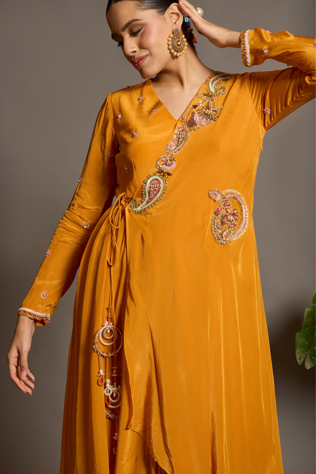 A vibrant marigold-orange Anarkali crafted in a fluid, lustrous fabric with a graceful layered silhouette. Adorned with intricate multicolour paisley embroidery and delicate scattered embellishments. Features a flattering tie-up waist with handcrafted tassels and elegant embroidered work. Shop Now!