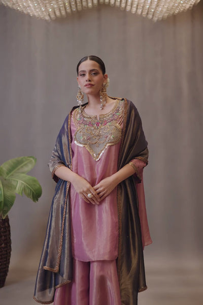 A luminous rose-pink kurta and flared palazzo set crafted in rich,metallic-finish fabric. Featuring an ornate multicolour embroidered neckline with intricate zardozi inspired detailing. Paired with a contrasting deep-blue dupatta finished with elegant gold borders. A festive ensemble that beautifully blends contemporary ease with traditional opulence. Shop Now!