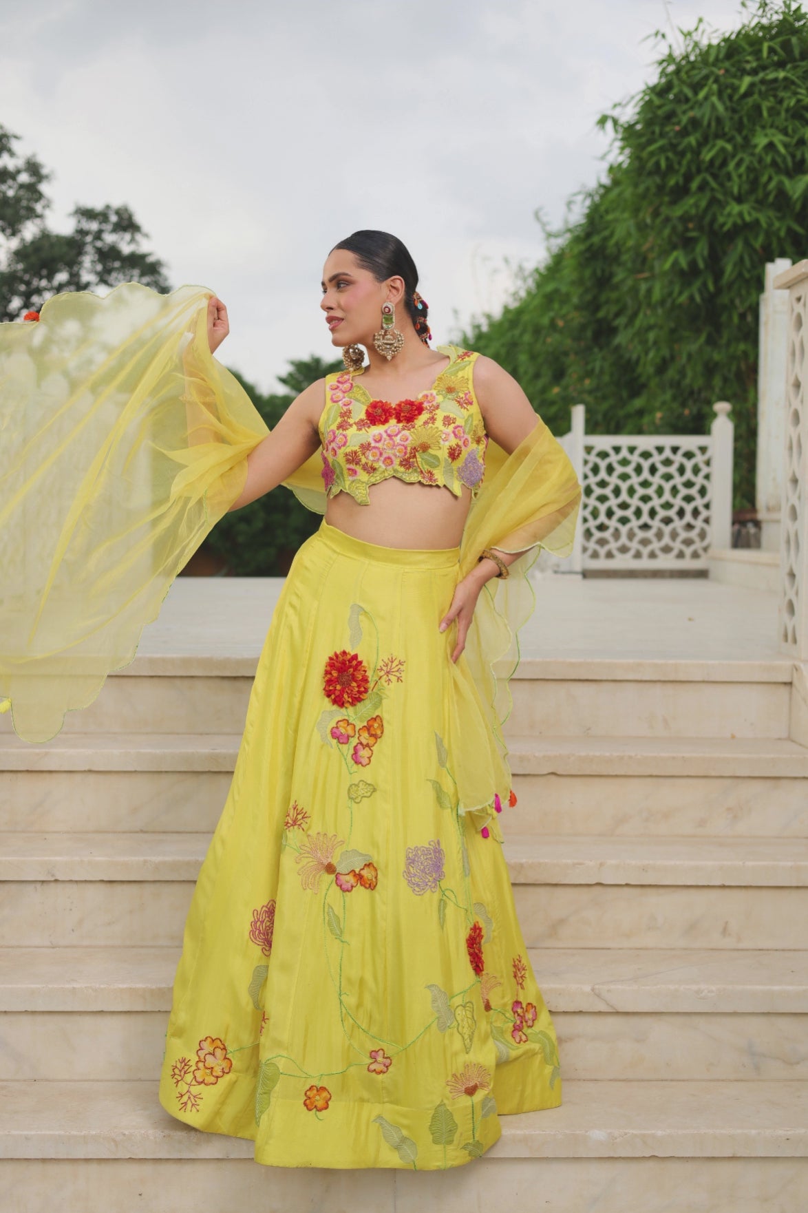 A dreamy sunshine-yellow lehenga set featuring a delicately embroidered floral appliqué blouse and flowing flared skirt. Intricate multicoloured botanical embroidery adds a fresh,playful charm, finished with a sheer scalloped dupatta and dainty tassel details. A vibrant, feminine festive ensemble that feels effortlessly desi,graceful and celebratory. Shop Now!