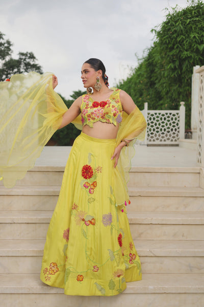 A dreamy sunshine-yellow lehenga set featuring a delicately embroidered floral appliqué blouse and flowing flared skirt. Intricate multicoloured botanical embroidery adds a fresh,playful charm, finished with a sheer scalloped dupatta and dainty tassel details. A vibrant, feminine festive ensemble that feels effortlessly desi,graceful and celebratory. Shop Now!